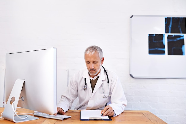 A doctor is sitting at his desk working on his computer and talking to a patient during a telemedicine call. The patient is an elderly woman and she is at home talking to the doctor in a virtual physical examination and is describing her symptoms to him with a smile.