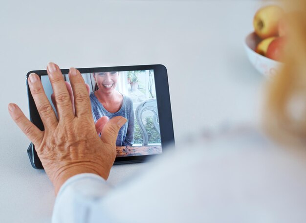 A close-up of a doctor holding a tablet during a video call with a patient. The doctor is smiling and talking, and the patient is visible on the screen. The background is blurred, indicating a clinical setting.